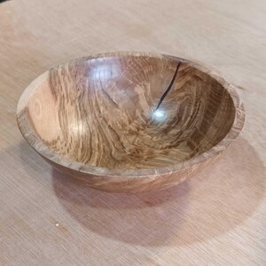 Może przedstawiać: Drewniana miska z naturalnym wykończeniem drewna. Miska ma jasnobrązowy kolor z ciemniejszymi brązowymi smugami w całym drewnie. Miska jest okrągła i ma gładką, wypolerowaną powierzchnię.