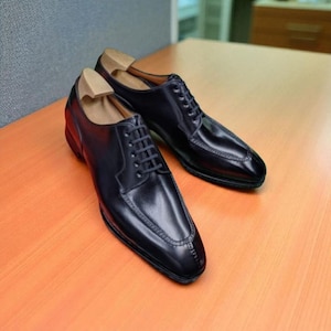 Peut inclure: Une paire de chaussures de ville en cuir noir au design classique. Les chaussures ont une finition brillante, une fermeture à lacets et des coutures décoratives. Des embauchoirs sont insérés. Les chaussures sont sur une surface en bois.