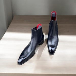 May include: A pair of black leather Chelsea boots with a glossy finish. The boots have a pointed toe and a red interior lining. The boots are displayed on a light-colored wooden surface.