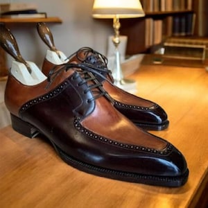 K&ouml;nnte beinhalten: Zweifarbige Lederschuhe in Braun und Schwarz. Die Schuhe haben ein klassisches Design mit spitzer Zehenpartie, schwarzen Schn&uuml;rsenkeln und dekorativen N&auml;hten. Schuhspanner sind eingesetzt. Die Schuhe stehen auf einer Holzoberfl&auml;che.