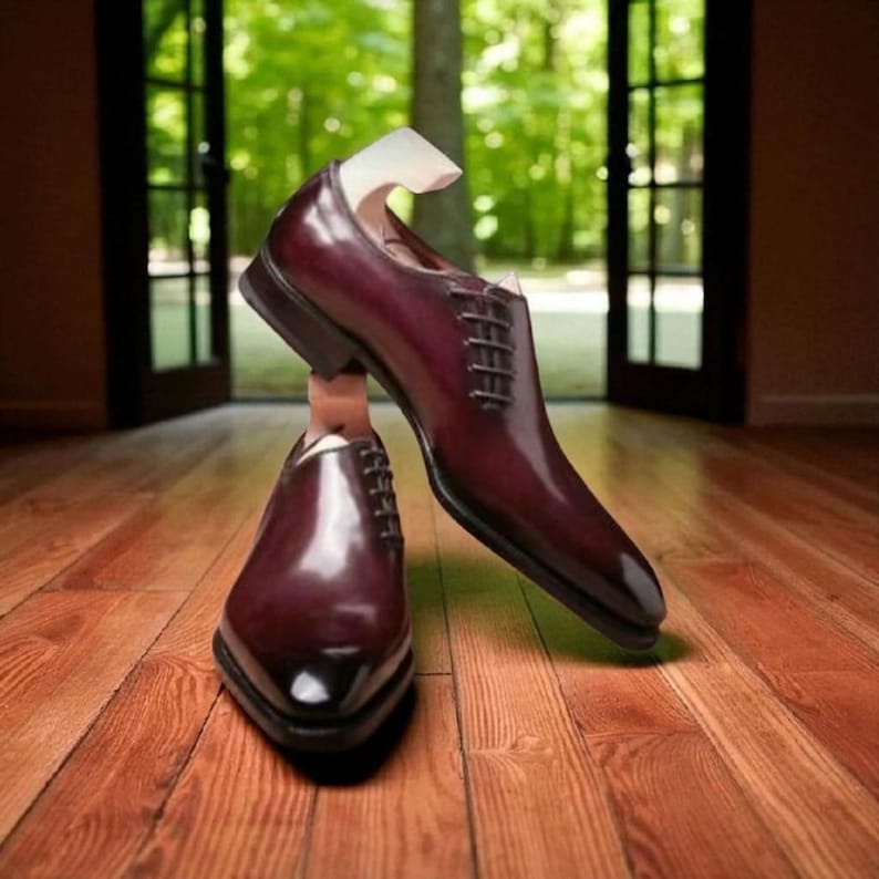 May include: A pair of burgundy leather dress shoes with a pointed toe and lace-up closure. The shoes are displayed with wooden shoe trees inside, showcasing their shape and design. The polished finish reflects light, highlighting the rich color.