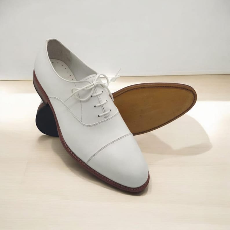 Pu&ograve; includere: Un paio di scarpe Oxford in pelle bianca dal design classico. Le scarpe hanno una punta arrotondata, una chiusura con lacci e una suola marrone. Le cuciture sono visibili e la suola &egrave; di un colore a contrasto. Le scarpe sono esposte su una superficie chiara.