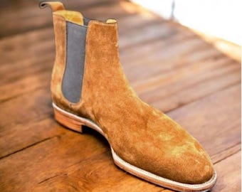May include: A tan suede Chelsea boot with a rounded toe and elastic side panels. The boot has a stacked heel and a pull tab at the back. The sole is made of a light-colored material. The boot is approximately 6 inches tall.