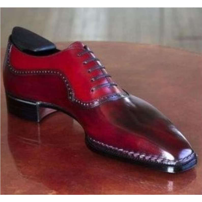 May include: A two-tone leather dress shoe in shades of red and burgundy. The shoe features a classic lace-up design with a pointed toe and decorative stitching along the edges. A black shoe tree is inserted inside the shoe.