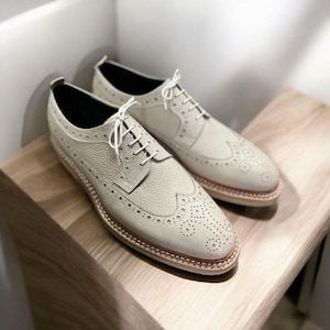 May include: A pair of off-white leather brogue shoes with white laces. The shoes have a wingtip design and a textured leather upper.