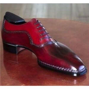 May include: A two-tone leather dress shoe in shades of red and burgundy. The shoe features a classic lace-up design with a pointed toe and decorative stitching along the edges. A black shoe tree is inserted inside the shoe.