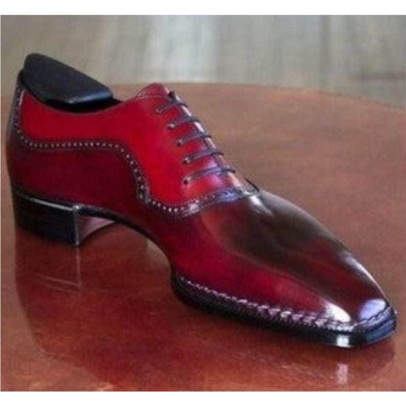 May include: A two-tone leather dress shoe in shades of red and burgundy. The shoe features a classic lace-up design with a pointed toe and decorative stitching along the edges. A black shoe tree is inserted inside the shoe.