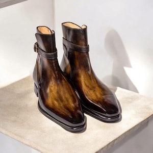 Puede incluir: Un par de botines de cuero marrón con un acabado pulido. Los botines tienen una correa con hebilla alrededor del tobillo y un tacón bajo. El cuero tiene un efecto degradado, pasando del marrón oscuro a un tono más claro.