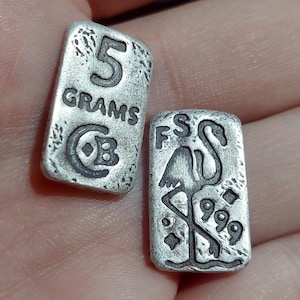 Pu&ograve; includere: Due piccole barre rettangolari d'argento. Una barra &egrave; stampata con "5 GRAMMI" e un simbolo. L'altra presenta un disegno di fenicottero con il testo "FS" e "999".