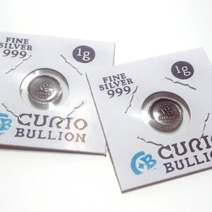 May include: Two silver 1g coins in protective packaging. The packaging is white with the text "FINE SILVER 999" and "CURIO BULLION". Each coin is visible through a circular window.