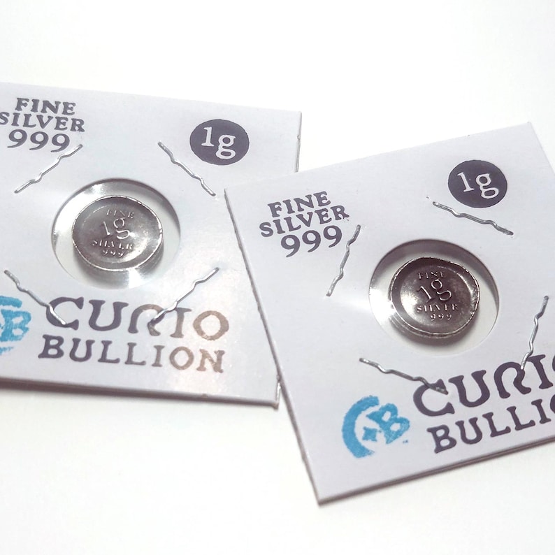 May include: Two silver 1g coins in protective packaging. The packaging is white with the text "FINE SILVER 999" and "CURIO BULLION". Each coin is visible through a circular window.