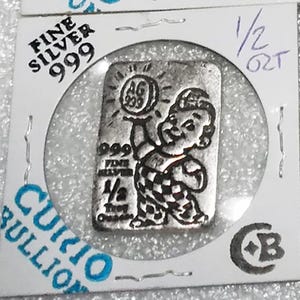 May include: A rectangular silver bullion bar weighing 1/2 troy ounce, encased in a white cardboard holder. The bar features an embossed cartoon figure holding a circular object marked AG 999, with text reading 999 FINE SILVER 1/2 Troy Ounce.