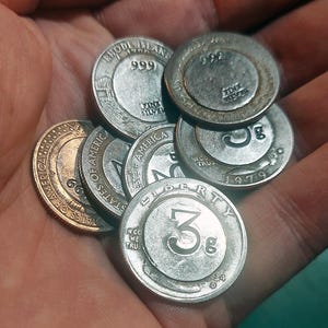 May include: A collection of circular silver-toned metal tokens and coins. Several pieces feature the text '3g' and 'Liberty', while others are marked 'Rhode Island', '999 fine silver', and 'States of America'. One coin displays the year 1979.