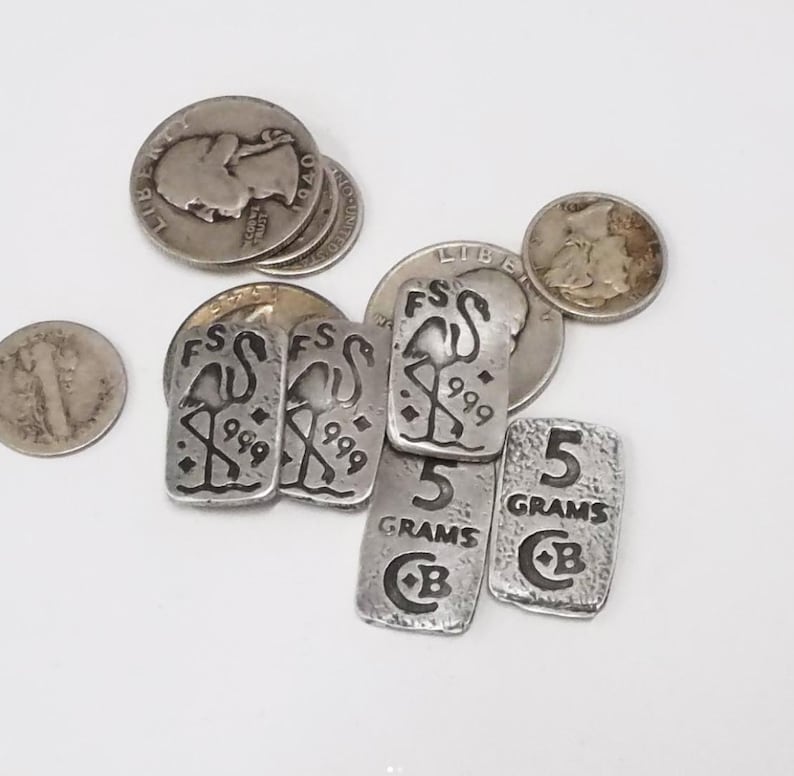 Pu&ograve; includere: Un assortimento di monete d'argento e lingotti rettangolari d'argento. I lingotti sono stampati con un disegno di fenicottero, il numero "999" e la scritta "5 GRAMMI". Le monete presentano vari disegni e testo.