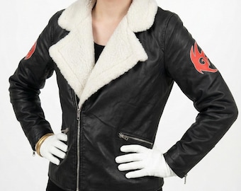 May include: A black leather-style jacket with a white faux-shearling collar, featuring red flame-like emblems on each sleeve. The jacket has a front zipper closure and zippered pockets. The outfit includes white gloves and a black and red wig with ears.