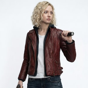 May include: A woman wearing a burgundy leather jacket over a white t-shirt and dark blue jeans. She is holding a flashlight in her right hand and a handgun in her left hand. The jacket has a zipper closure and multiple pockets.