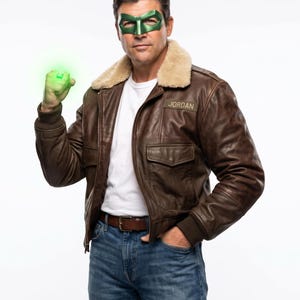 Puede incluir: Una chaqueta bomber de cuero marrón con cuello de piel sintética beige, una camiseta blanca y vaqueros azules. La chaqueta tiene un parche con el nombre JORDAN en el pecho. Se lleva un antifaz verde metálico y un anillo verde brillante.