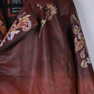May include: A brown leather jacket with decorative floral patterns in orange, yellow, and white. The jacket has a zipper closure and a label that reads "LEATHER JAC". The jacket is hanging on a black hanger.