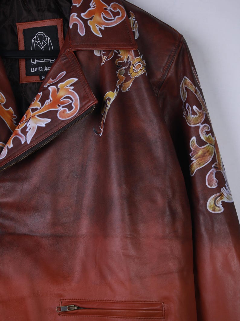 May include: A brown leather jacket with decorative floral patterns in orange, yellow, and white. The jacket has a zipper closure and a label that reads "LEATHER JAC". The jacket is hanging on a black hanger.