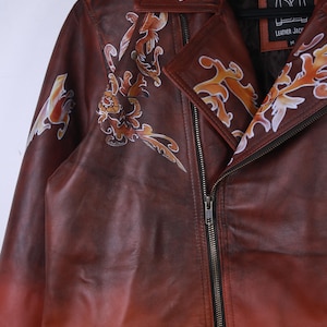 May include: A brown leather jacket with a gradient effect, featuring decorative floral designs in shades of orange and white. The jacket has a zipper closure and a collar. The tag reads "Leather Jacket".