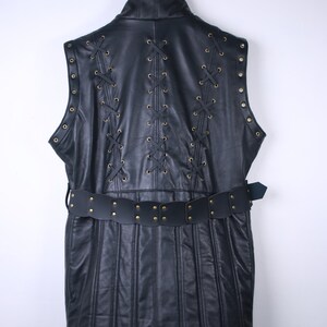 May include: Black leather vest with a high collar, featuring decorative lacing and brass-colored grommets. The vest has a belt with brass studs. The back of the vest has vertical stitching.