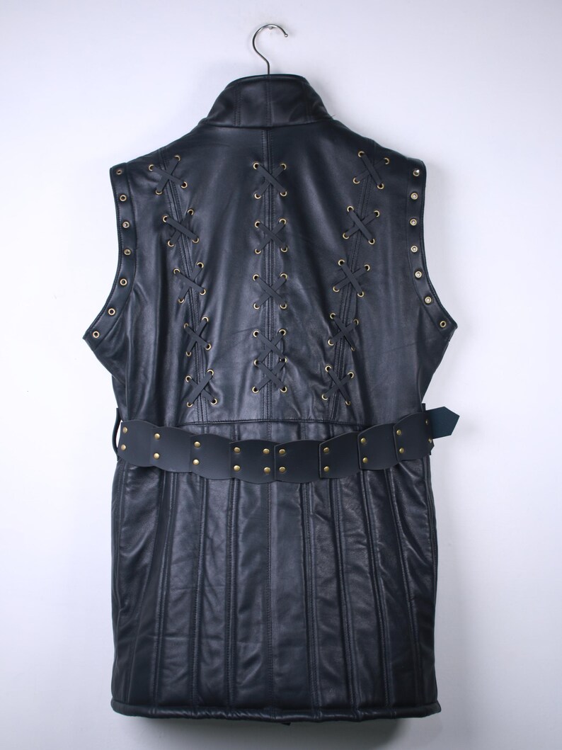 May include: Black leather vest with a high collar, featuring decorative lacing and brass-colored grommets. The vest has a belt with brass studs. The back of the vest has vertical stitching.