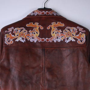 May include: Brown leather jacket with decorative orange and white floral design on the back. The jacket is hanging on a black hanger. The leather has a gradient effect, transitioning from brown to orange.