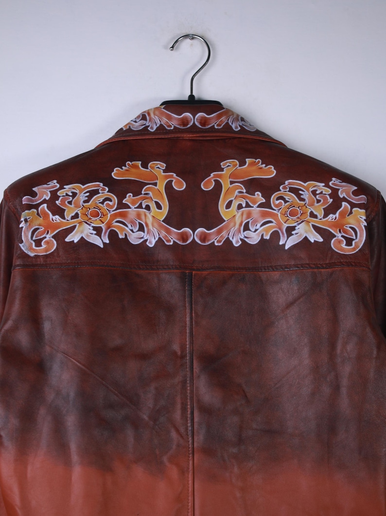 May include: Brown leather jacket with decorative orange and white floral design on the back. The jacket is hanging on a black hanger. The leather has a gradient effect, transitioning from brown to orange.