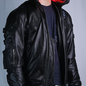 May include: A black leather jacket featuring a high collar with a glowing red light panel, a zippered utility pocket on the sleeve, and circular black accents. The jacket has a quilted front panel, ribbed cuffs, and a blue graphic detail on the shoulder.