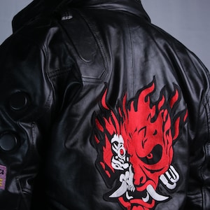 May include: A black leather jacket featuring a large red and white embroidered oni mask graphic on the back. The collar displays the white text SAMURAI. The jacket includes circular black accents on the sleeve and a small rectangular patch.