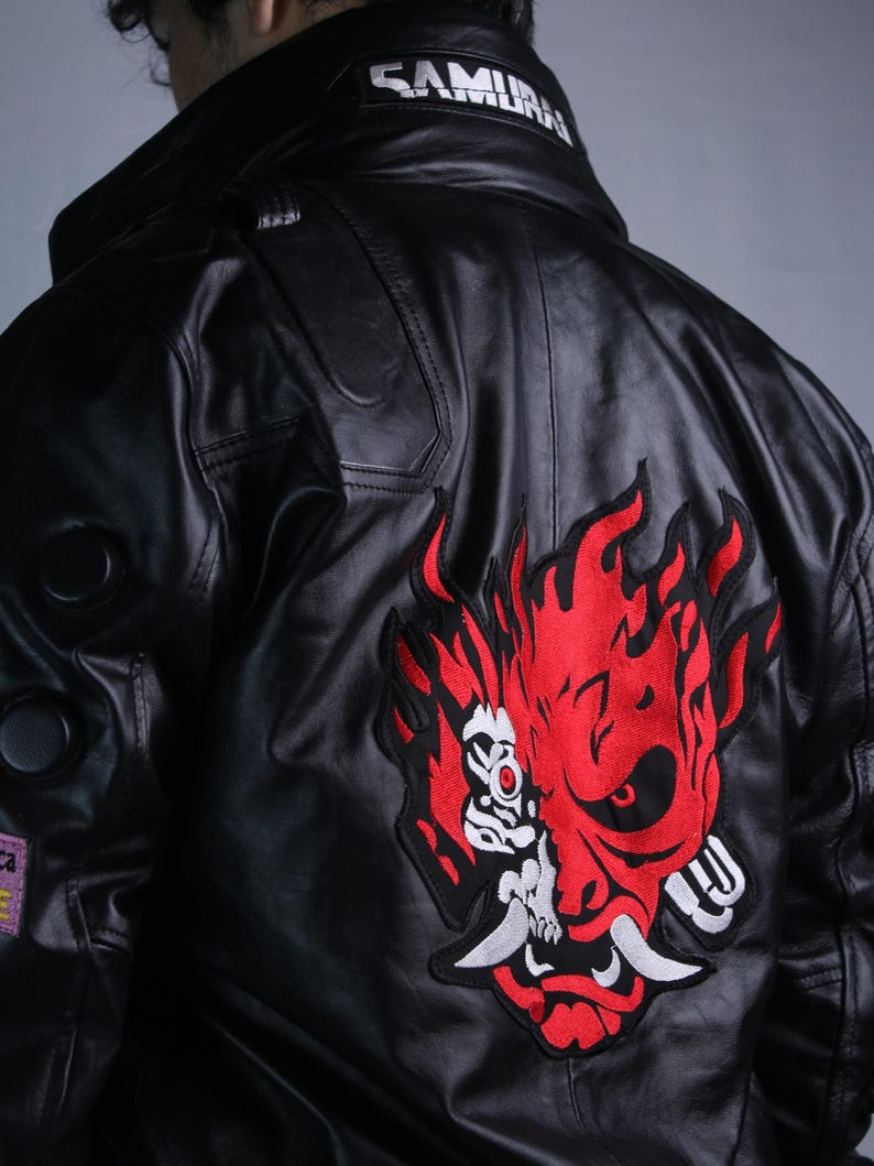 May include: A black leather jacket featuring a large red and white embroidered oni mask graphic on the back. The collar displays the white text SAMURAI. The jacket includes circular black accents on the sleeve and a small rectangular patch.