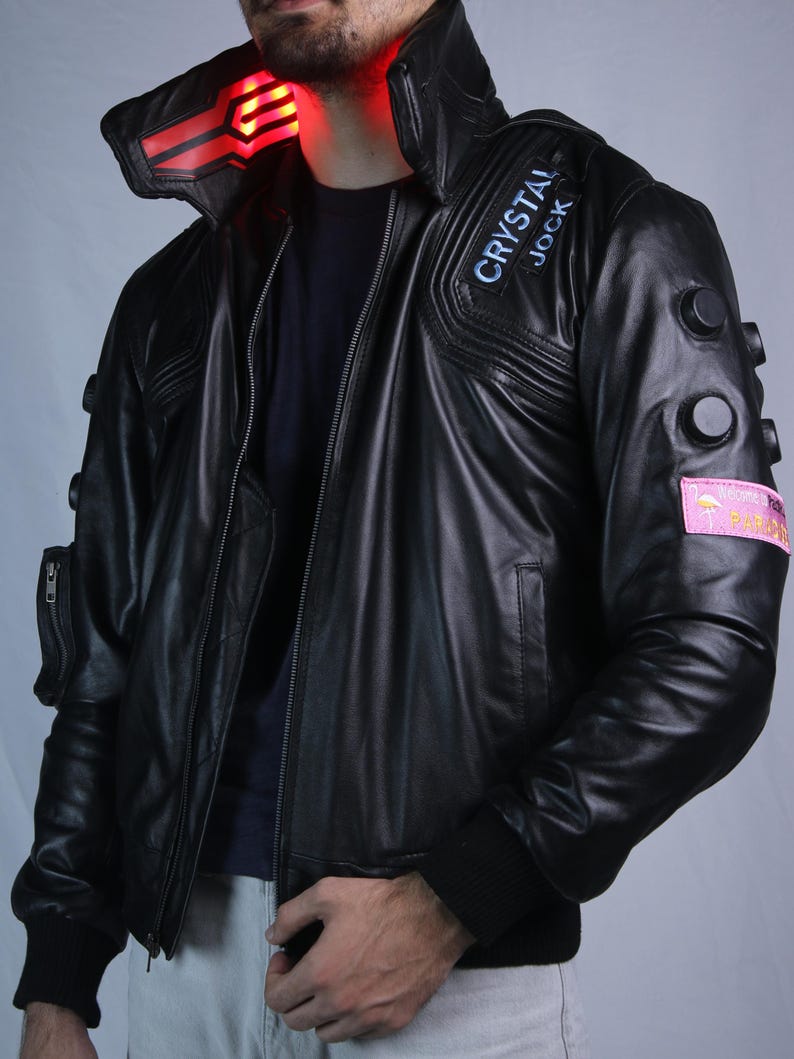 May include: A black leather jacket featuring a high collar with an illuminated red light panel, a front zipper, and black circular accents on the sleeves. A patch on the chest reads CRYSTAL JOCK, and a pink rectangular patch on the arm displays a flamingo.