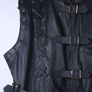 May include: Black leather vest with a high collar, featuring decorative lacing and brass-colored eyelets. The vest has three buckled straps and a belt with a buckle. The design is detailed and the leather appears to be of high quality.