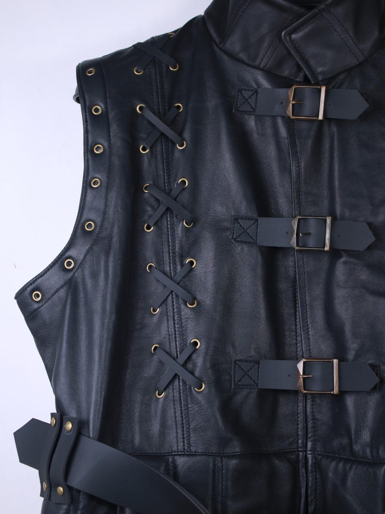 May include: Black leather vest with a high collar, featuring decorative lacing and brass-colored eyelets. The vest has three buckled straps and a belt with a buckle. The design is detailed and the leather appears to be of high quality.