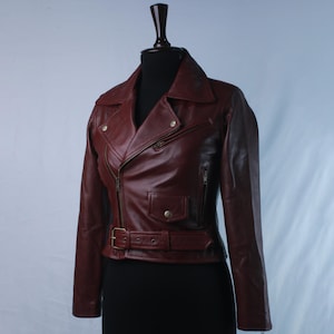 May include: A dark red leather motorcycle jacket featuring an asymmetrical front zipper, a notched lapel collar, a small flap pocket with a snap button, two zippered side pockets, and a buckled belt at the waist with metallic hardware.
