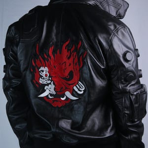 May include: A black leather jacket featuring a large red and white embroidered samurai mask graphic on the back. The collar displays the text SAMURAI in white lettering. The jacket includes circular black accents on the sleeves and a side pocket.
