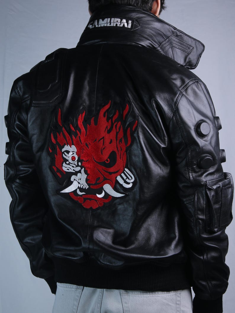 May include: A black leather jacket featuring a large red and white embroidered samurai mask graphic on the back. The collar displays the text SAMURAI in white lettering. The jacket includes circular black accents on the sleeves and a side pocket.