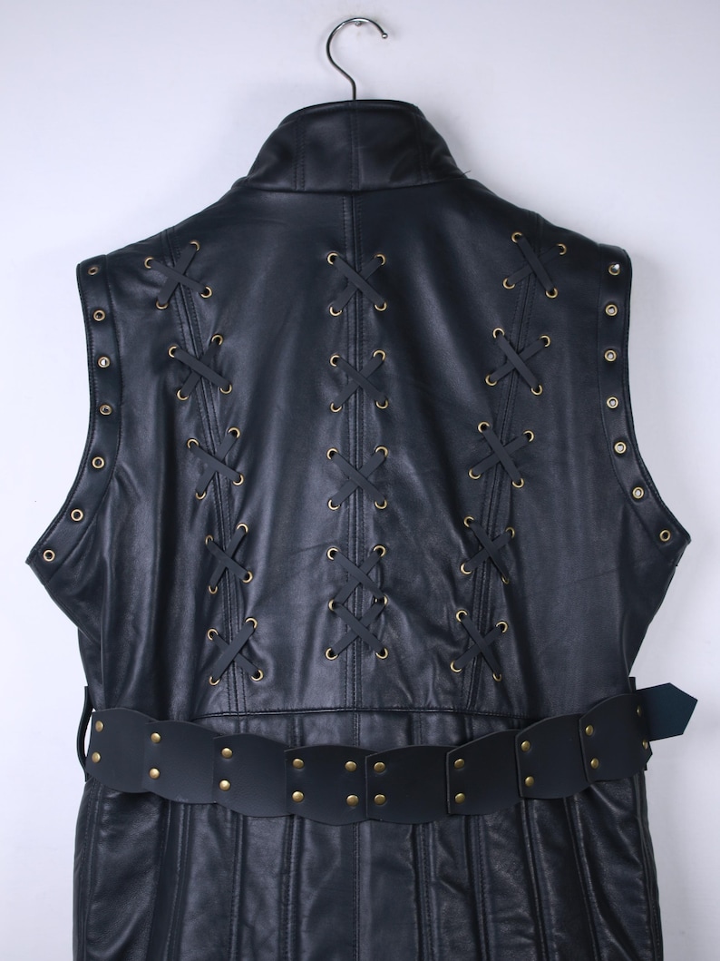 May include: Black leather vest with decorative lacing and a matching belt. The vest features brass-colored grommets and a high collar. The belt has brass-colored rivets. The vest is hanging on a metal hanger.