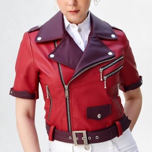 Puede incluir: Chaqueta de cuero roja con mangas cortas, cuello burdeos y cintur&oacute;n a juego. La chaqueta presenta cremalleras y botones plateados, y un peque&ntilde;o bolsillo burdeos. La persona lleva debajo una camisa blanca con cuello.
