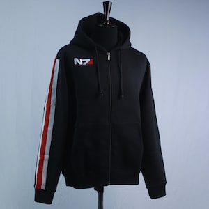 May include: A black zip-up hoodie featuring a hood with drawstrings and a full-length front zip. The chest displays the white and red N7 logo. The right sleeve is accented with a vertical stripe pattern in white, red, and white colours.