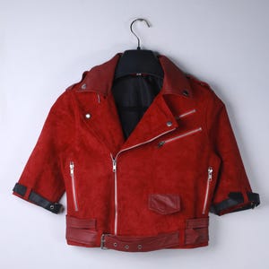 Puede incluir: Chaqueta de ante roja con cuello negro y mangas cortas. La chaqueta tiene cremalleras plateadas, cintur&oacute;n y pu&ntilde;os negros. La chaqueta est&aacute; colgada de una percha negra sobre un fondo blanco.