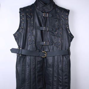 May include: Black leather vest with a high collar, metal grommets, and decorative lacing. The vest features multiple buckles and a belt at the waist. The garment is hanging on a black hanger.