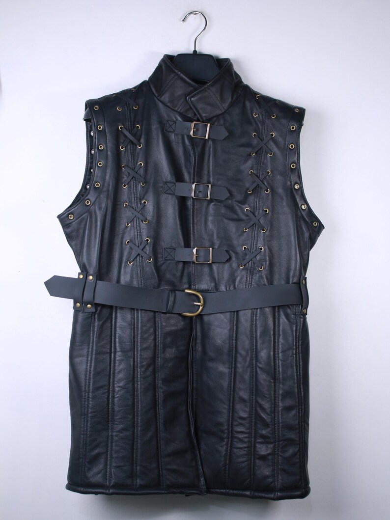 May include: Black leather vest with a high collar, metal grommets, and decorative lacing. The vest features multiple buckles and a belt at the waist. The garment is hanging on a black hanger.