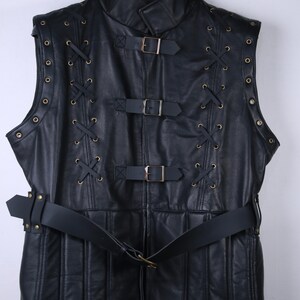 May include: A black leather vest with a high collar and a belted waist. The vest features decorative lacing, metal rivets, and three buckle closures. The garment is sleeveless and hangs from a black hanger.