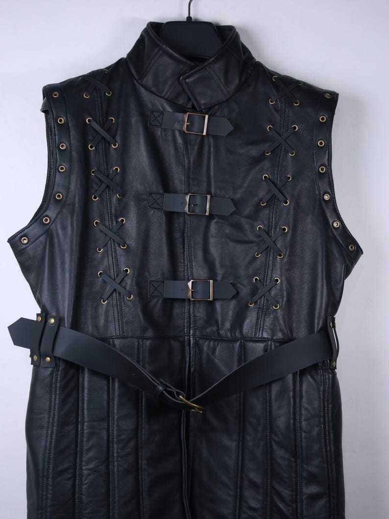 May include: A black leather vest with a high collar and a belted waist. The vest features decorative lacing, metal rivets, and three buckle closures. The garment is sleeveless and hangs from a black hanger.