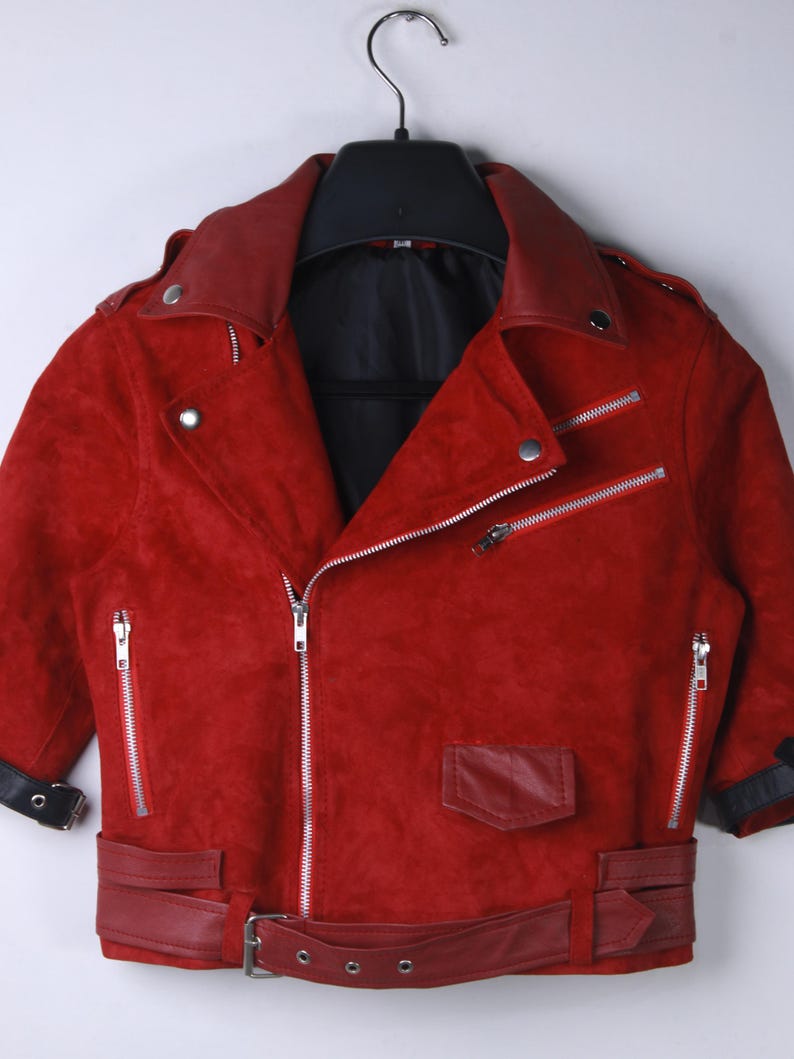 Puede incluir: Chaqueta roja de ante con estilo motero cl&aacute;sico. Cuenta con cuello con solapas, m&uacute;ltiples cremalleras plateadas, un peque&ntilde;o bolsillo y cintur&oacute;n en la cintura. La chaqueta est&aacute; colgada en una percha negra.