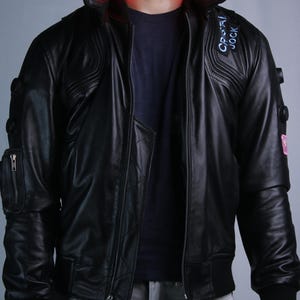 May include: A black leather jacket featuring a high collar with integrated red LED lighting, a front zipper closure, and a patch on the chest with the text "CRISTAL JOCK". The sleeves include circular accents and a zippered utility pocket.