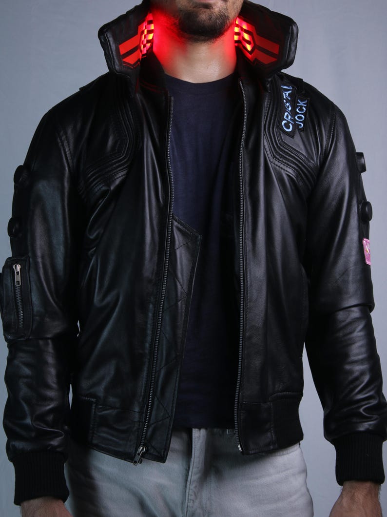 May include: A black leather jacket featuring a high collar with integrated red LED lighting, a front zipper closure, and a patch on the chest with the text "CRISTAL JOCK". The sleeves include circular accents and a zippered utility pocket.