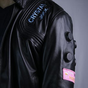 May include: A black leather jacket featuring a shoulder patch with the text CRYSTAL JOCK in blue lettering. The sleeve includes three circular black studs and a rectangular pink patch displaying a flamingo graphic and the text PARADISE.