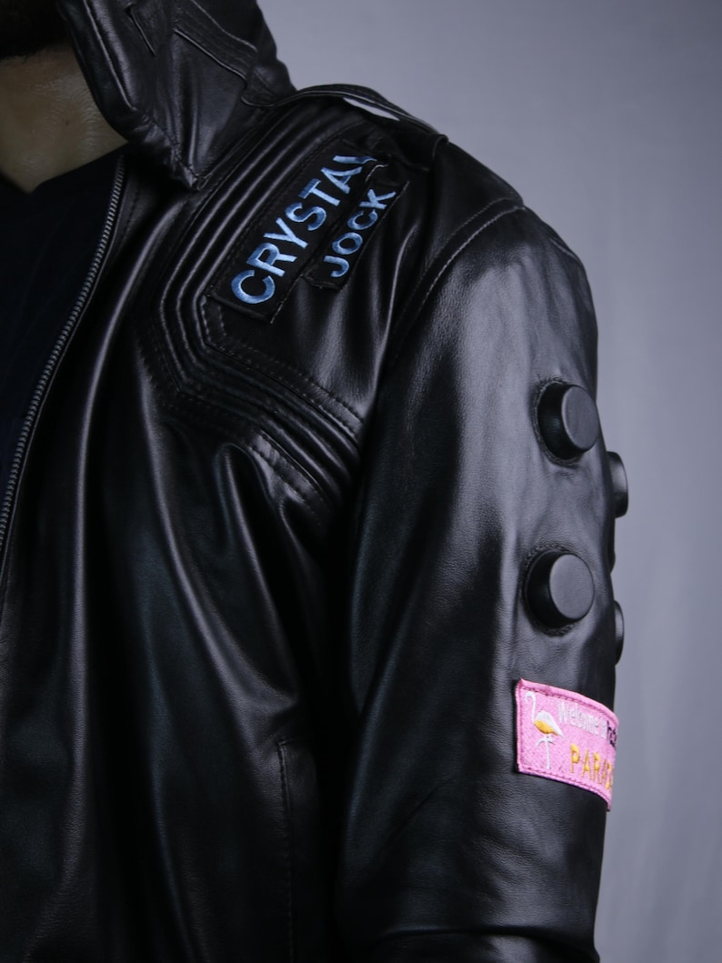 May include: A black leather jacket featuring a shoulder patch with the text CRYSTAL JOCK in blue lettering. The sleeve includes three circular black studs and a rectangular pink patch displaying a flamingo graphic and the text PARADISE.